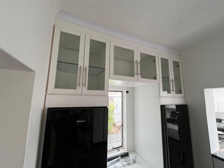 White wall cupboards with glass doors and glass shelves, built up to the cornice over two black fridges